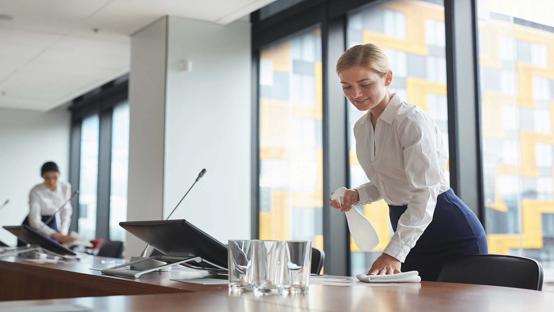 Portrait of smiling young woman cleaning table with sanitizing spray in conference room while preparing for business event in office, copy space.