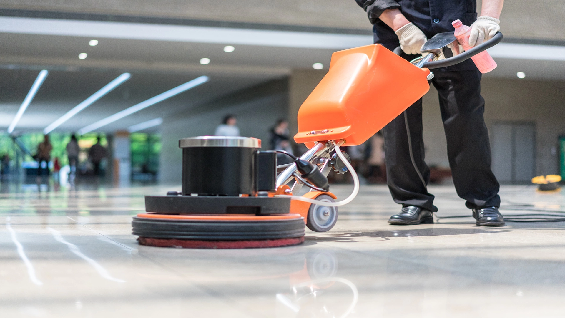 Person operating an orange and black floor cleaning machine in a large indoor space with tiled floor, pillars, and people walking in the distance, with a row of windows on the top allowing natural light to enter.