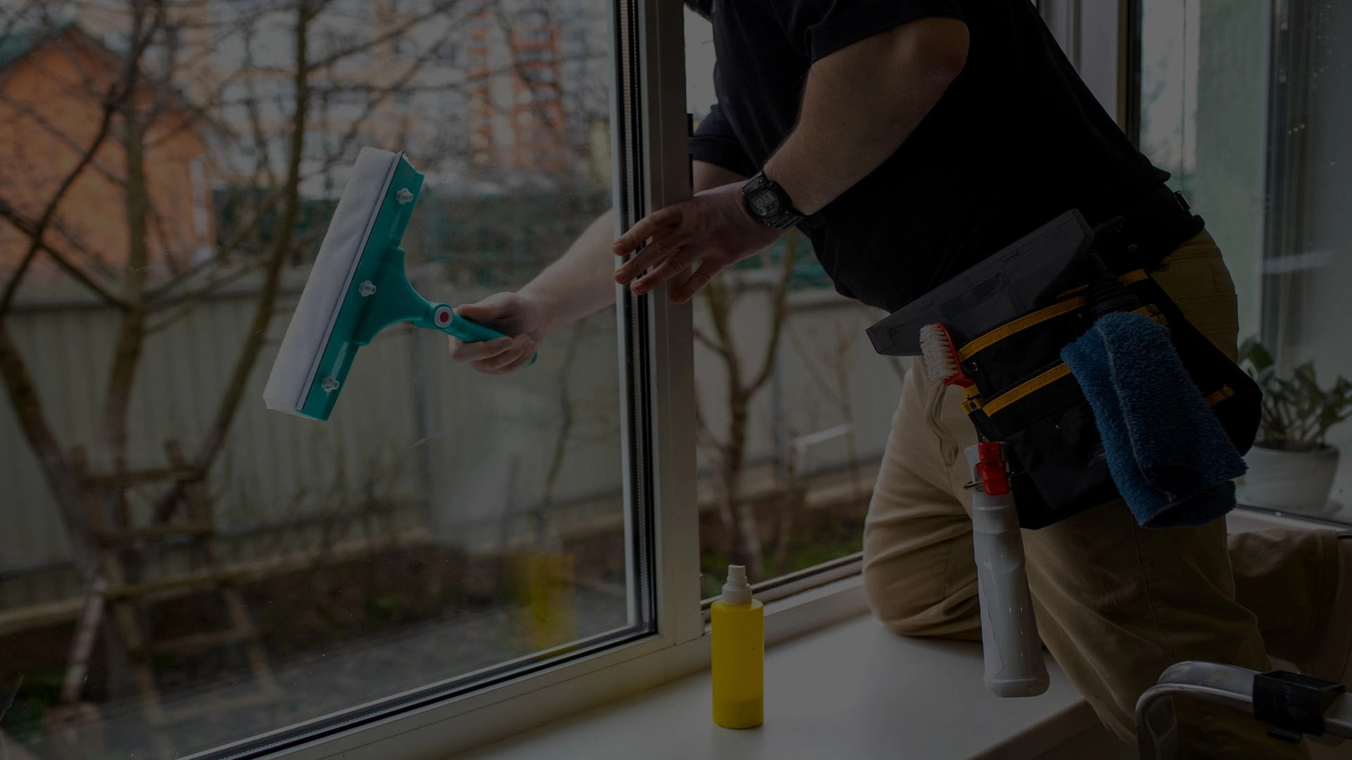 Person cleaning a large window with a turquoise squeegee, wearing a black t-shirt and beige pants.