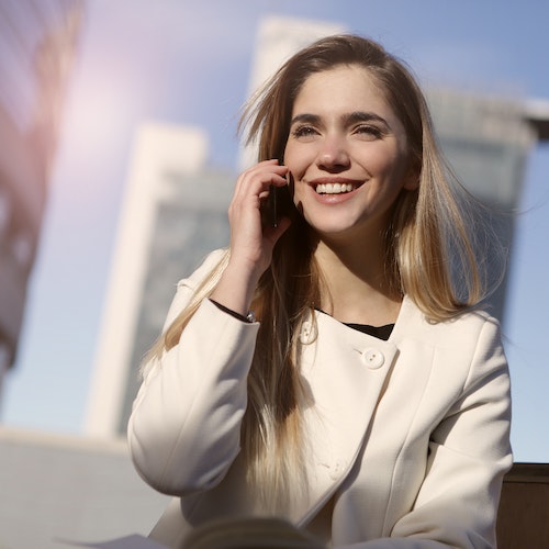 A girl in a white coat sitting outside with a city skyline in the background, holding a phone to their ear during the day with clear blue skies.