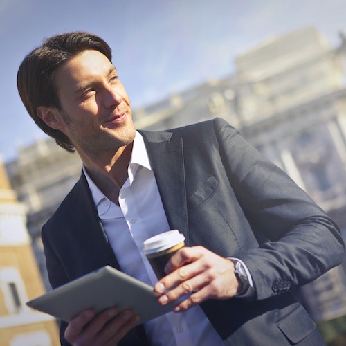 A man in a black suit and white shirt holding a coffee cup and a tablet, standing in front of a cityscape with buildings and blue sky.