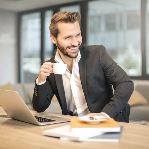 A person in a black suit and white shirt sitting at a desk in an office setting, holding a white coffee cup in their right hand with a laptop and a notebook on the desk, and a window with a view of a building and a plant on the windowsill in the background.