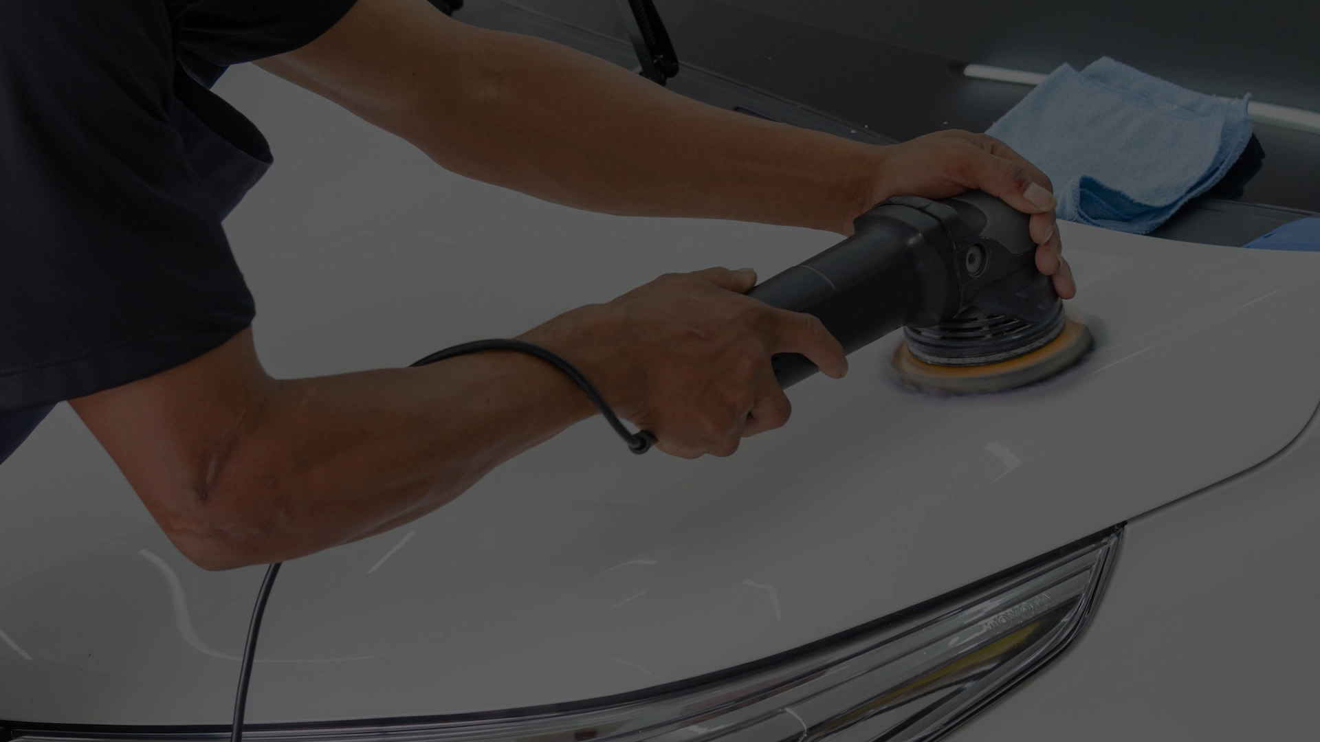 A hand holding a black and yellow polisher on the hood of a white car in a garage or workshop.