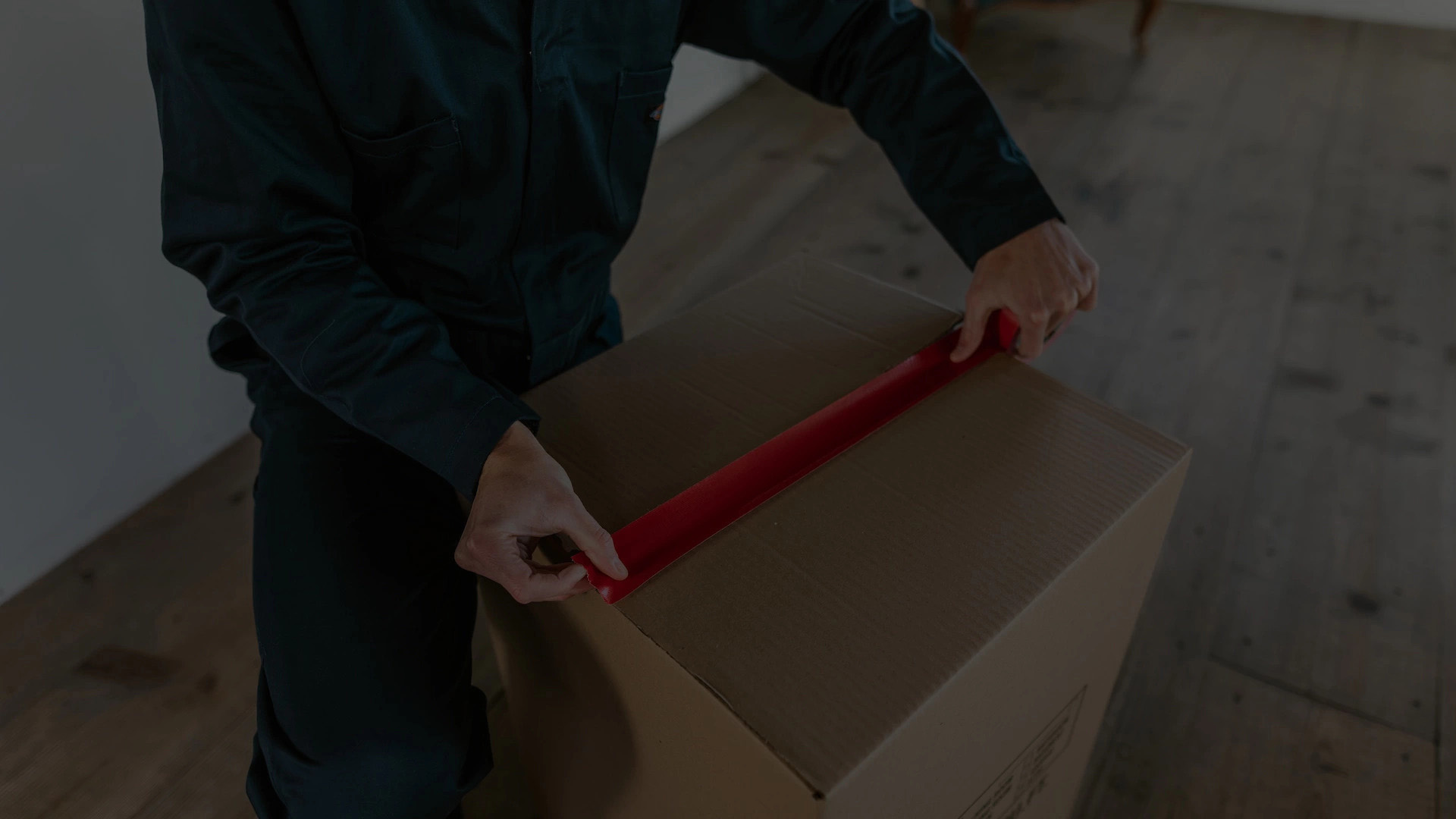 A person in a green jacket using a red tape dispenser to seal a large cardboard box on a wooden floor.