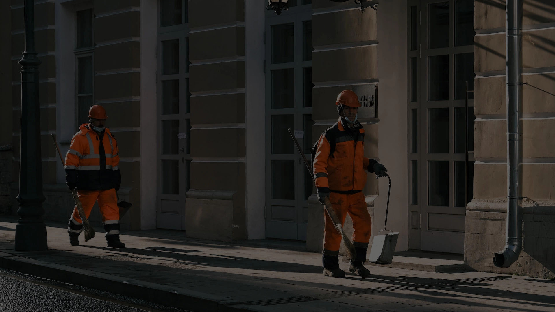 Two construction workers in orange vests and hard hats walking on a sidewalk in front of a white building.