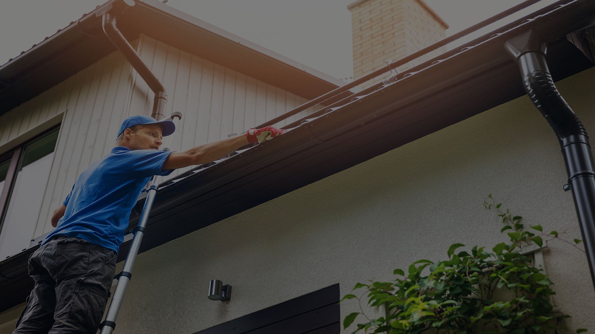 Man cleaning gutters on white house with brown roof. Wearing blue shirt, cap, gray pants. Silver ladder, blue sky.